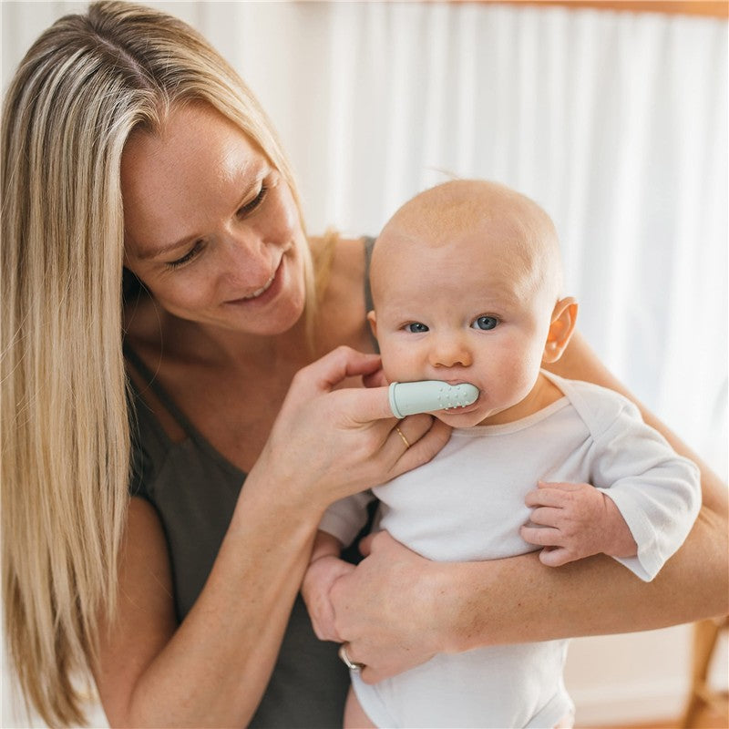 A woman is gently using a gray and light green silicone finger toothbrush on a baby in her arms, promoting dental hygiene.