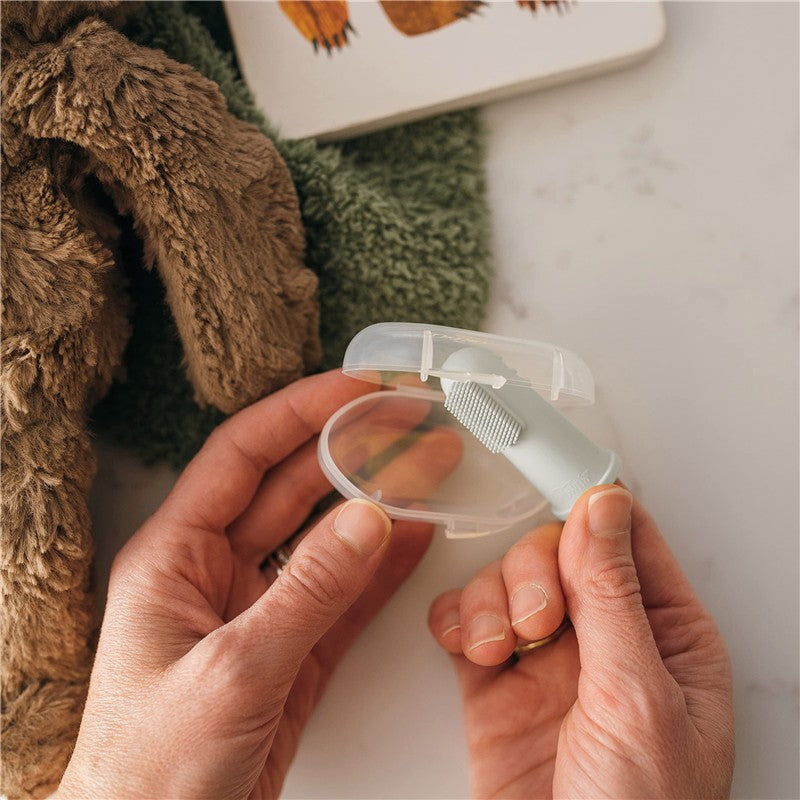 Silicone finger toothbrush in a clear case, held by a hand, with a plush toy and green cloth in the background.