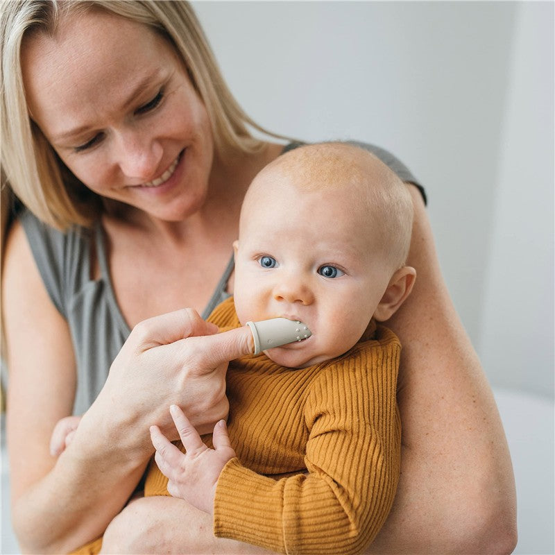 A mother is smiling while using a gray silicone finger toothbrush on her baby, who is wearing an orange ribbed shirt.