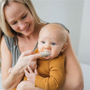 A mother is smiling while using a gray silicone finger toothbrush on her baby, who is wearing an orange ribbed shirt.