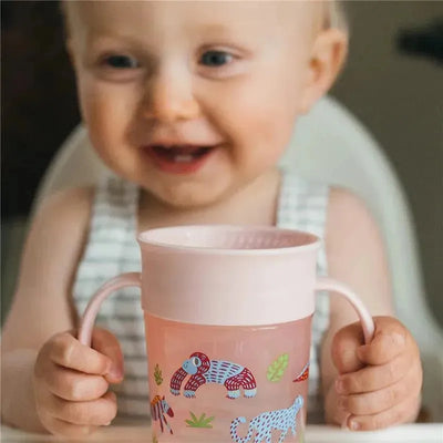 Baby smiling while holding a pink Dr. Brown Cheers360 cup with colorful animal designs.