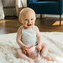 Smiling baby sitting on a soft rug, holding a pink Dr. Brown Cheers360 Cup with colorful designs.