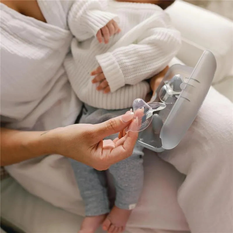 Clear nipple shields held by a caregiver, with a baby resting in their lap, and a sterilizer case nearby.