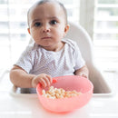 Baby sitting in a high chair, holding a pink bowl filled with puffed snacks, looking curiously at the camera.