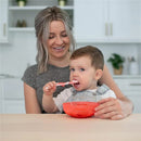 A toddler eating from a bright red bowl while holding a toothbrush, seated with a smiling adult.