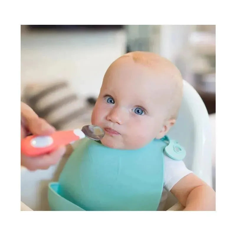 A baby wearing a blue bib sits in a high chair, looking at a spoon with a coral handle being offered to them.