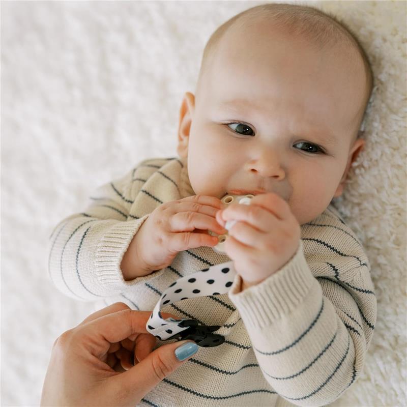Baby holding a pacifier, with a polka-dotted clip being attached to their clothing.