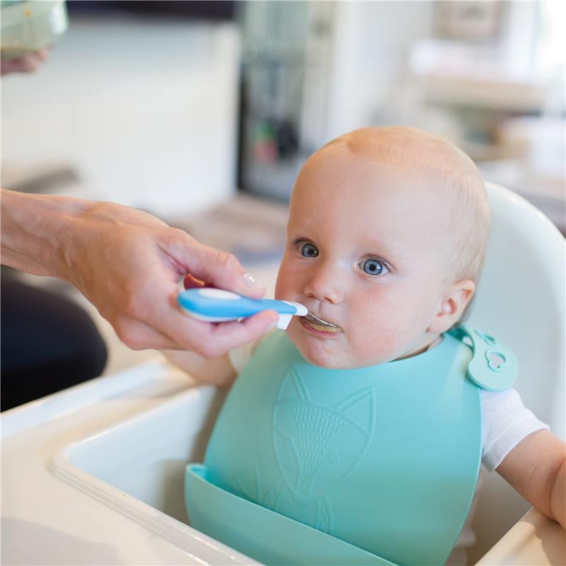Toddler with blue bib being fed with a soft-grip spoon by an adult in a bright kitchen setting.