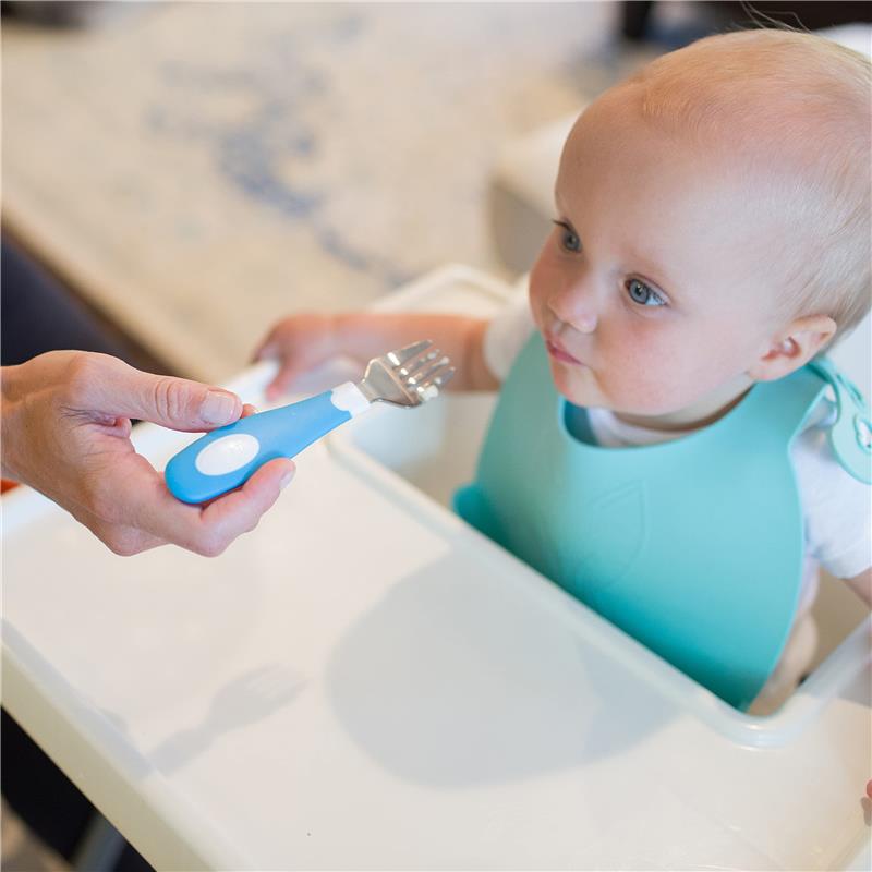 A toddler in a blue bib looks curiously at a blue soft-grip fork being offered by an adult.