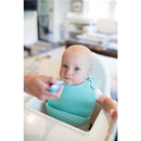 Toddler in a high chair wearing a light blue bib, being fed with a soft-grip spoon by an adult.