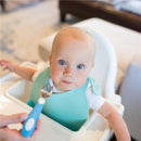 Toddler in a high chair with a blue soft-grip fork, wearing a bib, looking at the camera.