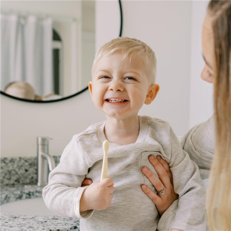 Smiling toddler holding a toothbrush, wearing a gray shirt, while being held by an adult in a bathroom setting.