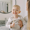 Smiling toddler holding a toothbrush, wearing a gray shirt, while being held by an adult in a bathroom setting.