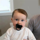 A baby wearing a white sweater holds a black pacifier, gazing at the camera with big blue eyes.