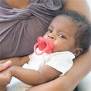 Infant in white onesie holding a pink pacifier, being cradled by an adult wearing a grey top.