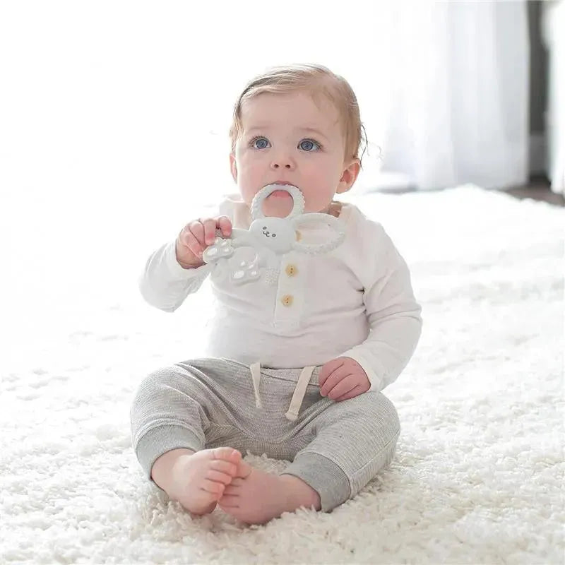Gray silicone teether shaped like a bunny held by a baby sitting on a soft rug, wearing a white long-sleeve shirt and gray pants.