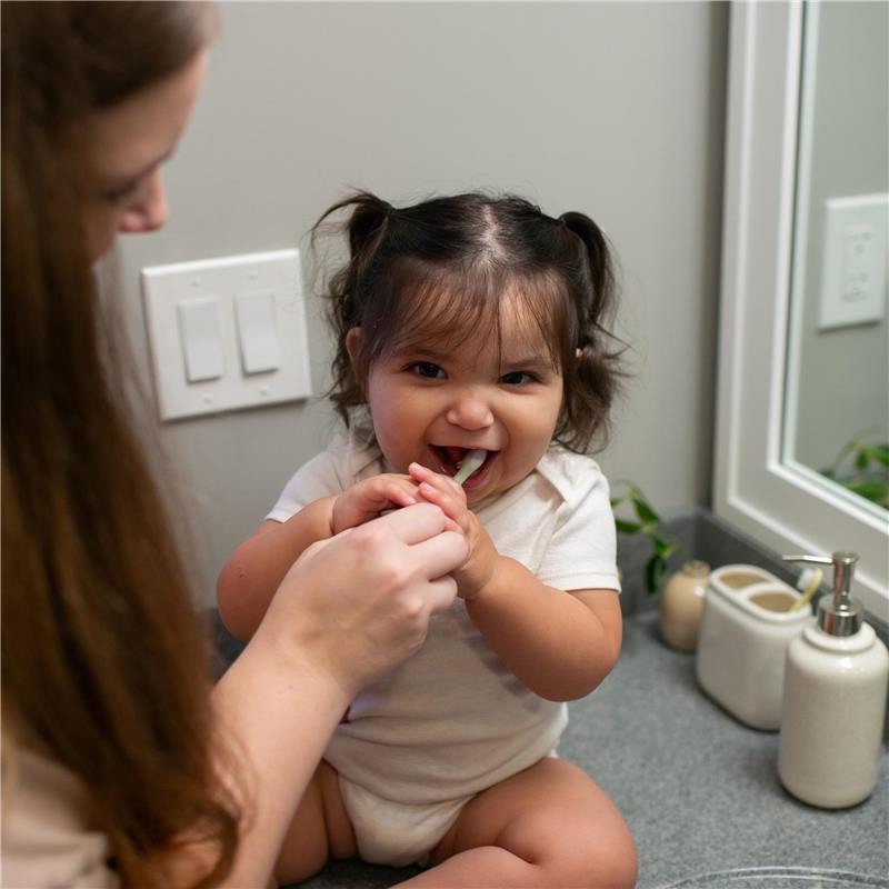 Toddler with pigtails smiling while brushing teeth with an elephant-themed toothbrush, assisted by an adult.