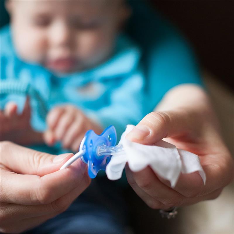 A caregiver uses a wipe to clean a blue pacifier while a baby is partially visible in the background.