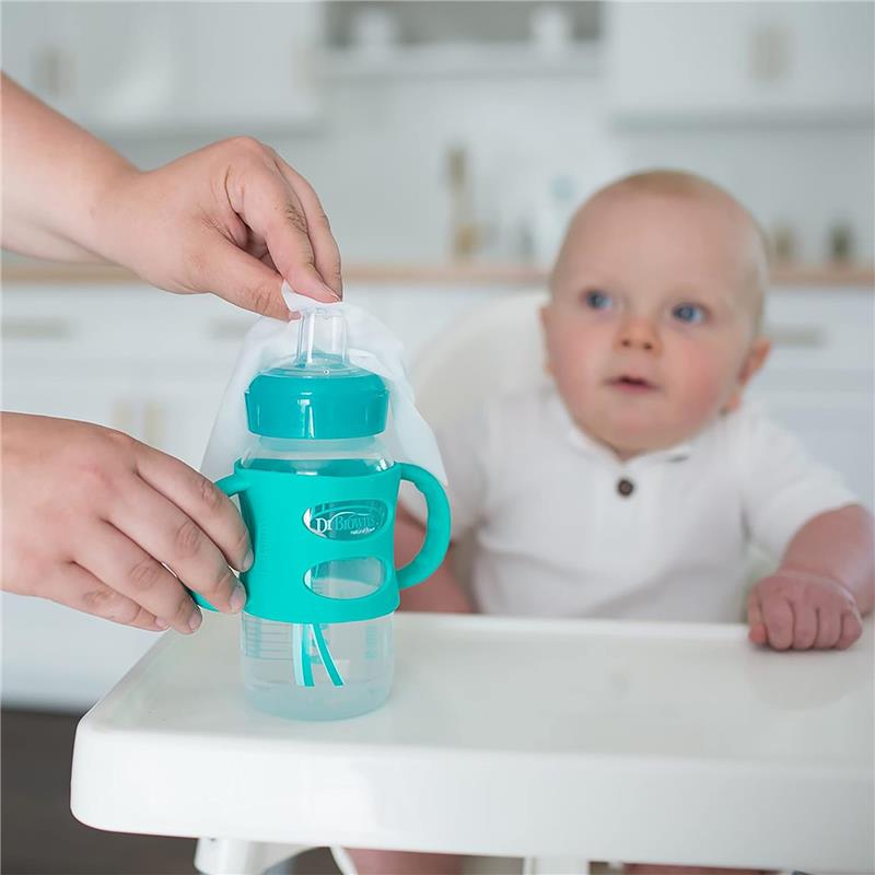 Hand cleaning a teal Dr. Brown sippy cup with a wipe, while a baby observes curiously from a high chair.