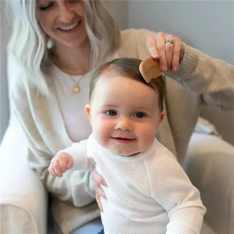 A smiling baby in a white sweater is being brushed with a wooden comb by a woman in a cozy setting.