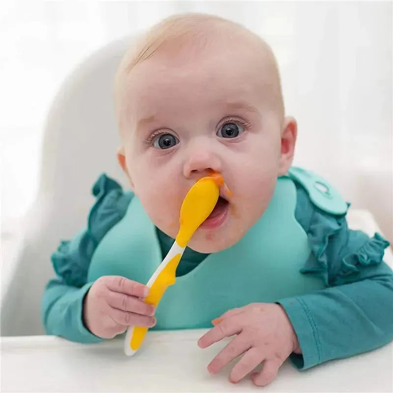 A baby with blue eyes holds a yellow spoon, with food smeared on their face and a teal bib on.