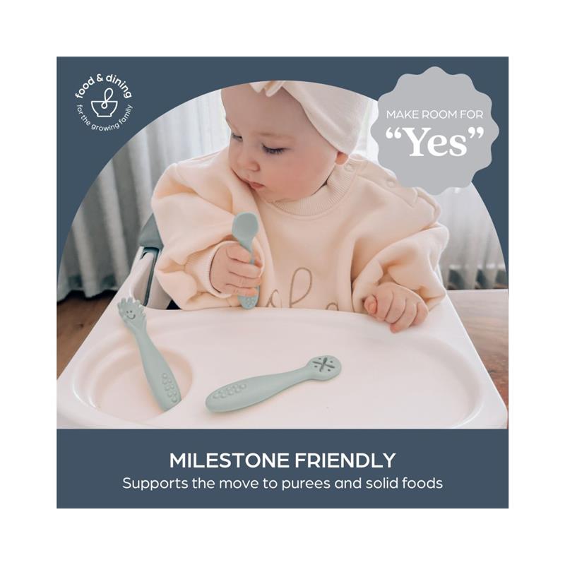A baby wearing a cream sweater and headband holds a spoon, with two seafoam green utensils on a high chair tray.