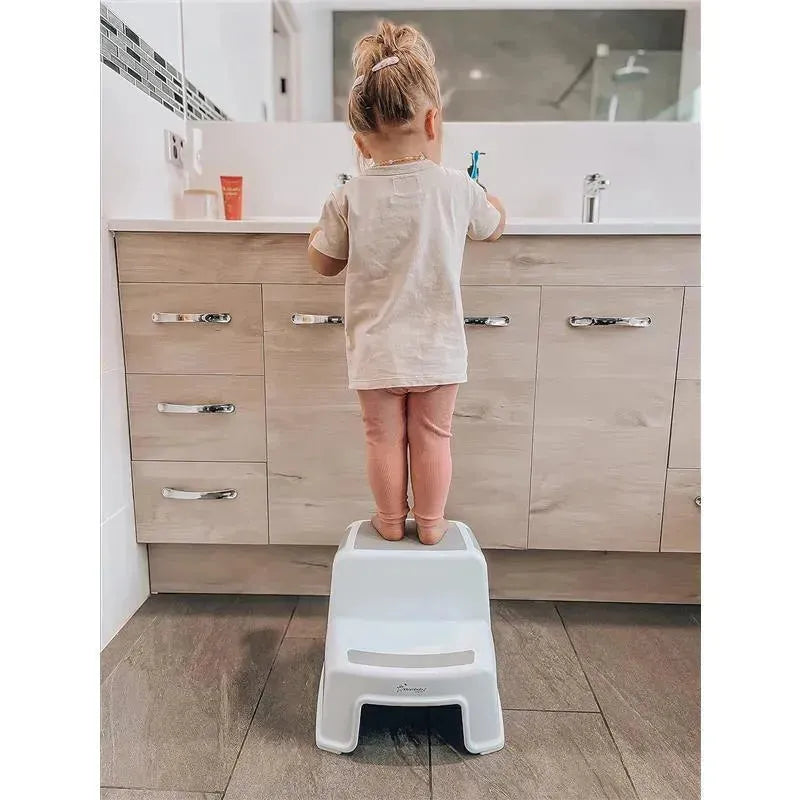 A toddler standing on a grey and white Dreambaby 2-Up step stool at a bathroom sink, brushing their teeth.
