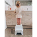 A toddler standing on a grey and white Dreambaby 2-Up step stool at a bathroom sink, brushing their teeth.