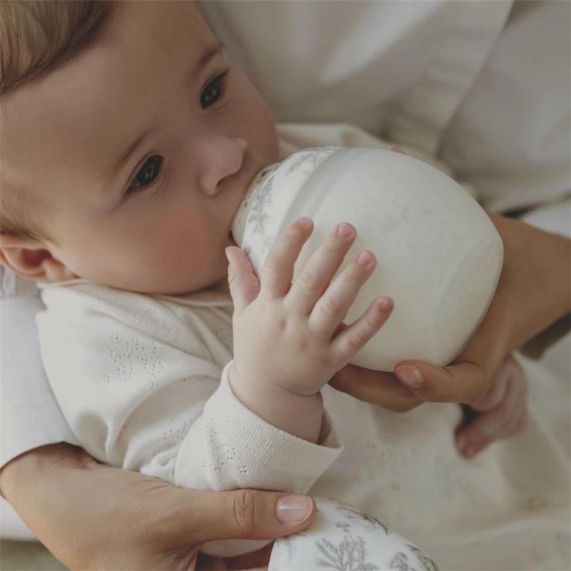 Baby drinking from a decorative grey Elhee bottle, held by a caregiver, featuring soft focus and warm tones.
