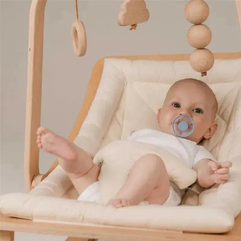 Baby in a light-colored baby seat with a pacifier, surrounded by wooden mobile toys, appearing calm and relaxed.