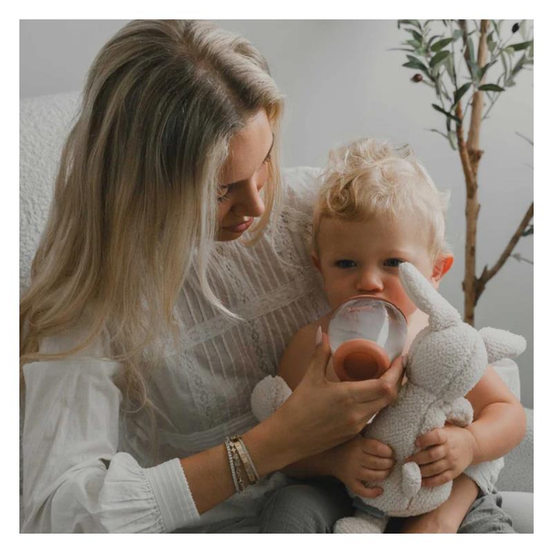 Mother feeding toddler from a glass bottle with a deep nipple, while the child holds a soft toy bunny.
