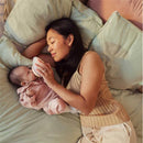 A woman gently feeds a baby with a bottle while lying on a bed surrounded by soft pillows.