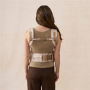 Ergobaby Alta Hip Seat Baby Carrier in natural beige, shown on a woman from the back, featuring padded straps and adjustable buckles.