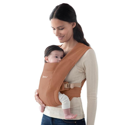 Ergobaby Embrace Newborn Baby Carrier in Canyon Clay, worn by a woman carrying a baby snugly against her chest.