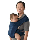 Ergobaby Embrace newborn baby carrier in navy, worn by a woman holding an infant close to her chest.