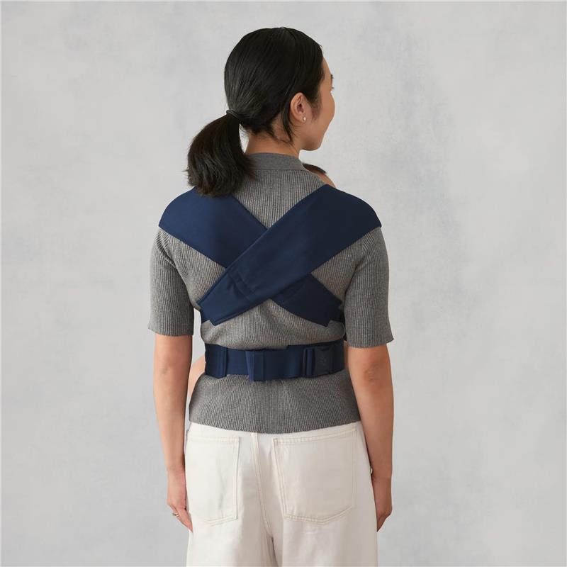 Ergobaby Embrace Newborn Baby Carrier shown worn on a woman, featuring crossed shoulder straps and a secure waist belt.