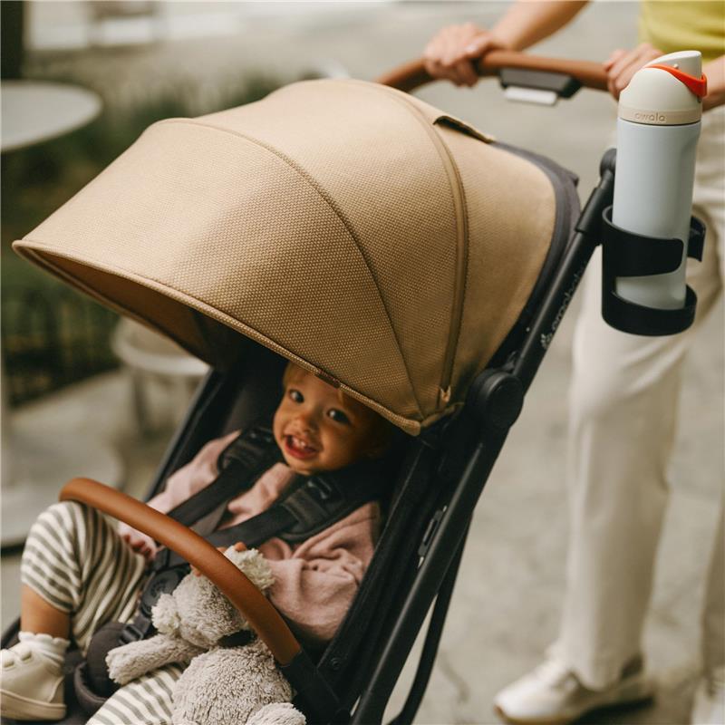 Ergobaby Metro 3 Deluxe Baby Stroller in camel color, featuring a smiling baby holding a plush toy under a large canopy.