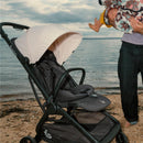 Ergobaby Metro 3 stroller on a beach, showcasing a white canopy, adjustable seat, and a parent holding a child nearby.