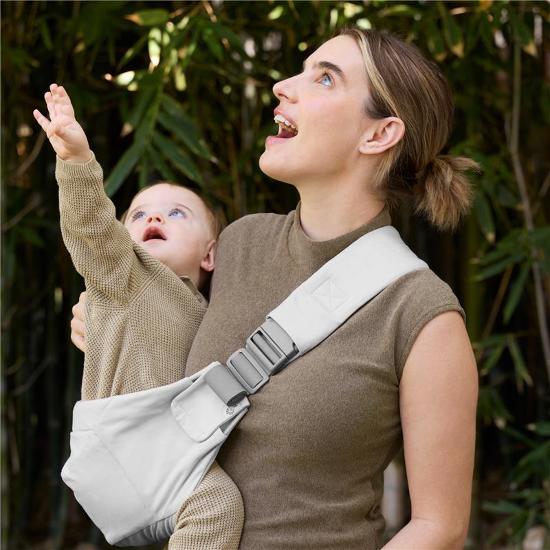 A woman wearing a pearl grey Ergobaby Upsie Sling holds a toddler who reaches upward, both smiling, with greenery in the background.