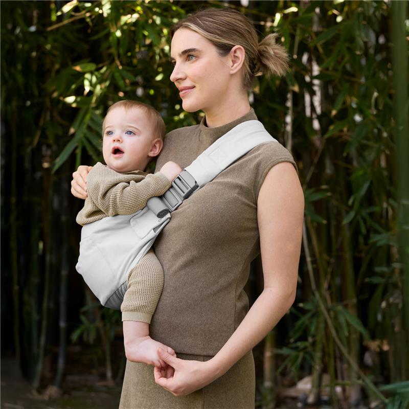 Mother wearing an Ergobaby Upsie Sling Baby Carrier in Pearl Grey, holding a toddler against her side in a bamboo setting.