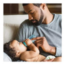A father feeds a baby with a turquoise Evenflo Feeding plastic bottle while sitting on a couch.