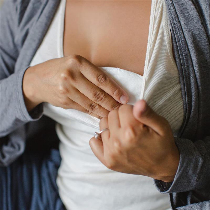 A woman adjusts a nursing pad underneath her clothing while wearing a light gray cardigan, demonstrating use of Evenflo Feeding Advanced Nursing Pads.