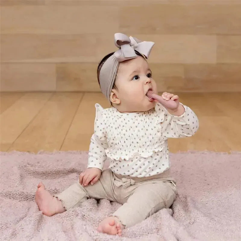 A baby sitting on a soft rug, holding and chewing a blush-colored baby toothbrush, wearing a floral shirt and headband.
