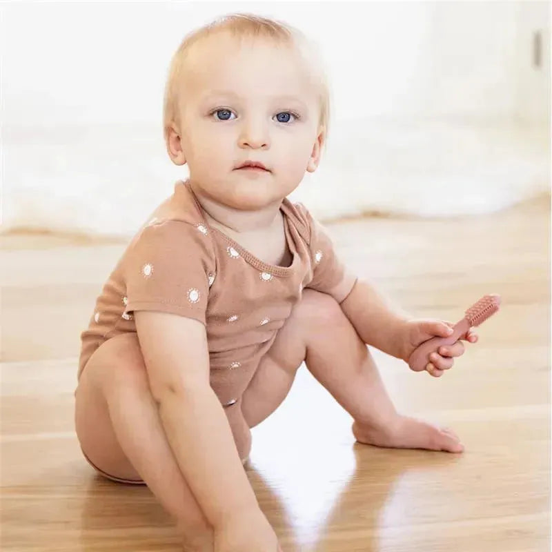 A baby sitting on the floor, holding a blush-colored Ezpz Baby-Led Toothbrush, wearing a light brown onesie with white dots.