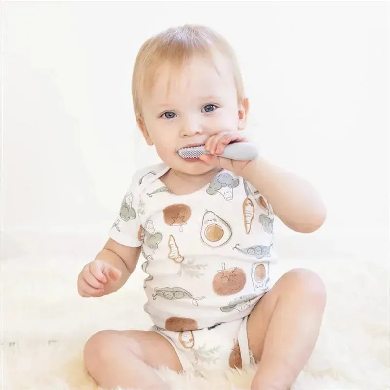 Toddler in a patterned onesie using the Ezpz Baby-Led™ Toothbrush in pewter color to brush their teeth.