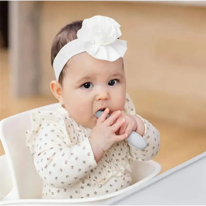 Baby in a high chair, wearing a headband with a flower, using an Ezpz Baby-Led Toothbrush in pewter color.