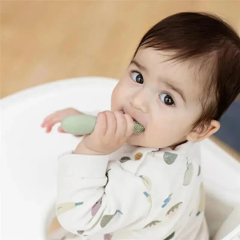 A baby with dark hair and a patterned onesie holds a sage green Baby-Led™ Toothbrush, exploring oral care.