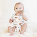 A toddler wearing a food-patterned onesie holds a sage-colored, baby-led toothbrush, focusing on brushing.