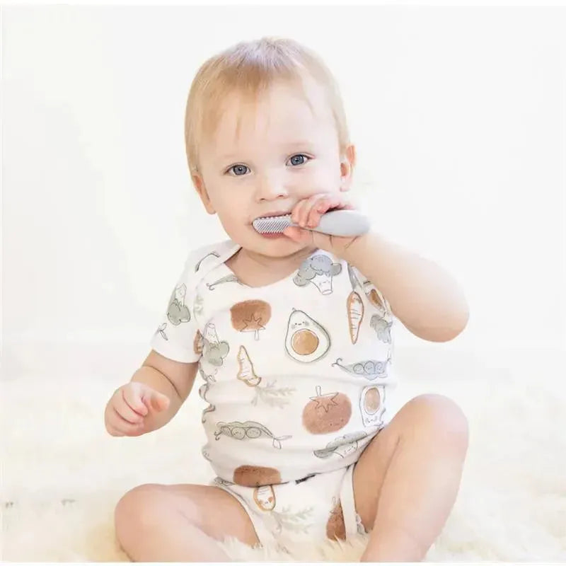 Toddler using a gray Ezpz Baby-Led toothbrush while sitting on a soft surface, wearing a patterned onesie.