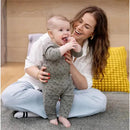 A smiling baby in a gray outfit playing with a toothbrush while being held by a parent, with a cozy background.